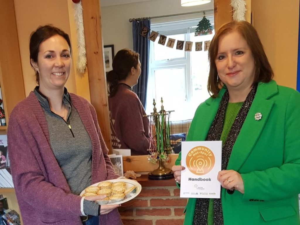 Two women stand by a fireplace. One holds a plate of minced pies and the other (Lauren fron Norfolk Community Foundation) holds a booklet.