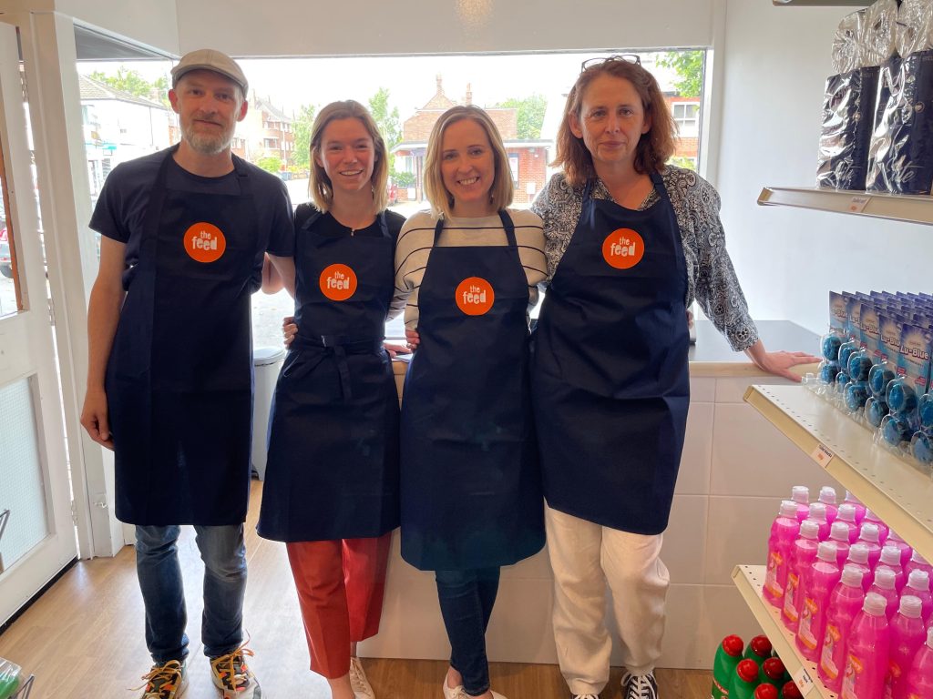 Volunteers and staff at The Feed in Norwich. They stand in a line in front of the till, wearing matching blue aprons with the Feed's orange logo on them.