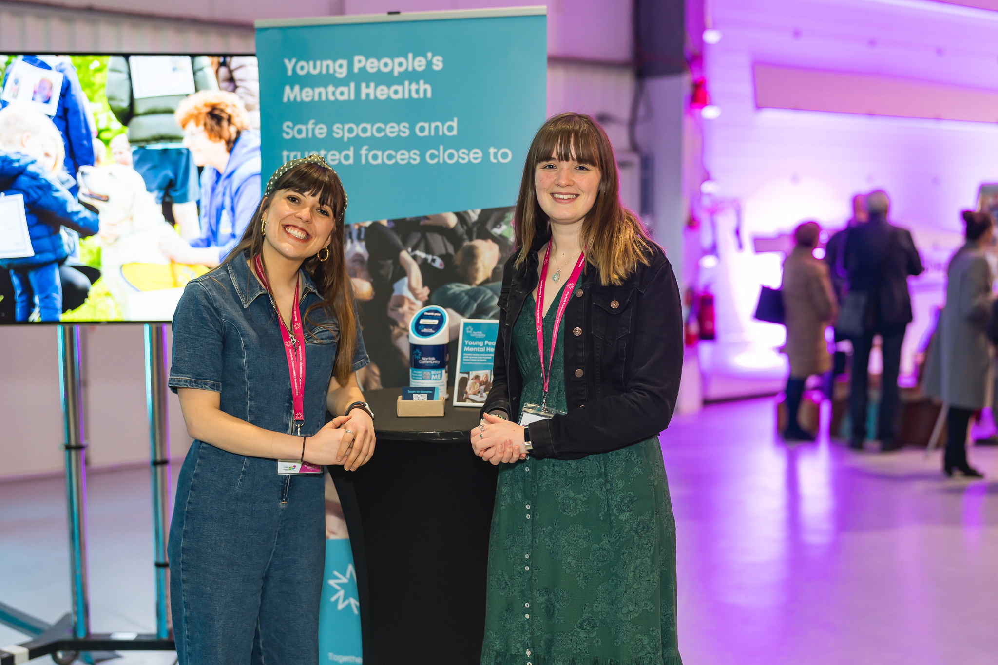Two women stand leaning on a tall black table. Behind them is a banner talking about Young People's Mental Health.